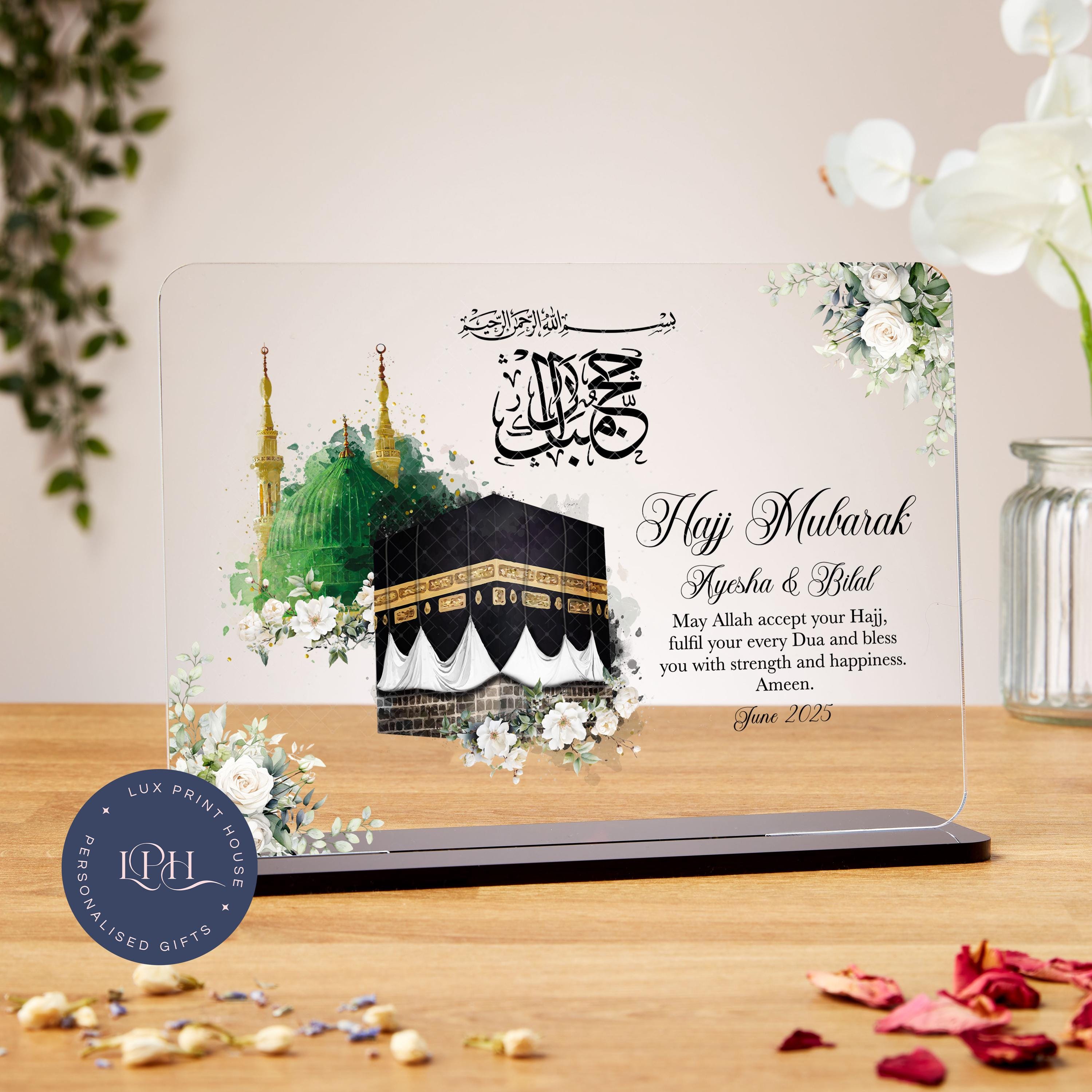 A personalised Hajj Mubarak acrylic plaque featuring detailed illustrations of the Kaaba and the Green Dome of Madinah, adorned with soft white floral accents. Arabic calligraphy reads “Hajj Mubarak,” accompanied by a dua asking Allah to accept the Hajj, fulfil every dua, and bless with strength and happiness. Includes space for custom names and date, making it a meaningful keepsake for commemorating this sacred journey.