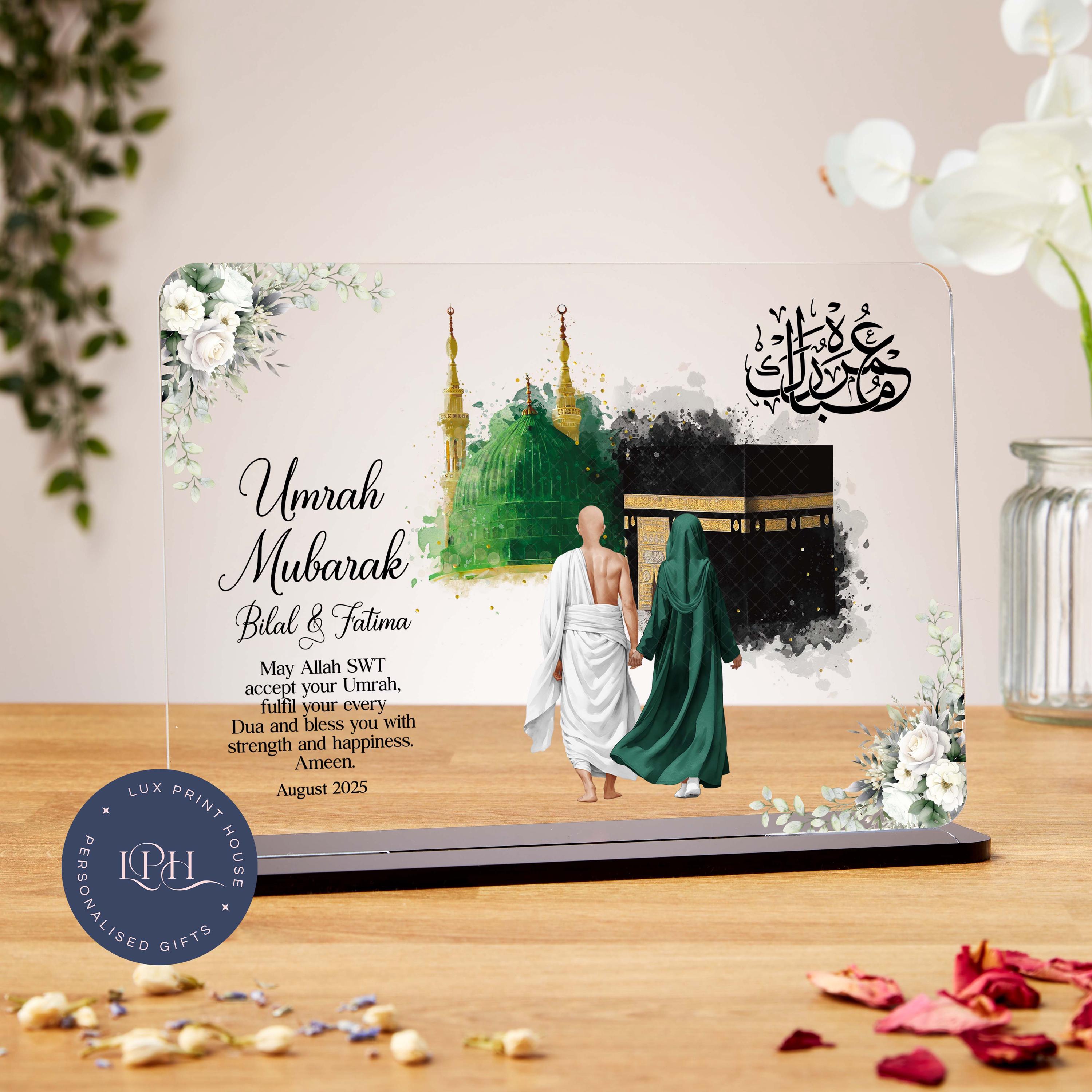 A personalised Umrah gift plaque featuring a faceless illustration of a couple holding hands, dressed in traditional Umrah attire, walking toward the Kaaba. The Green Dome of Madinah is also illustrated in the background. Surrounded by soft white floral accents and Arabic calligraphy that reads “Umrah Mubarak,” the design includes a dua asking Allah to accept the Umrah and bless the recipients with strength and happiness. Customisable name and date fields included.