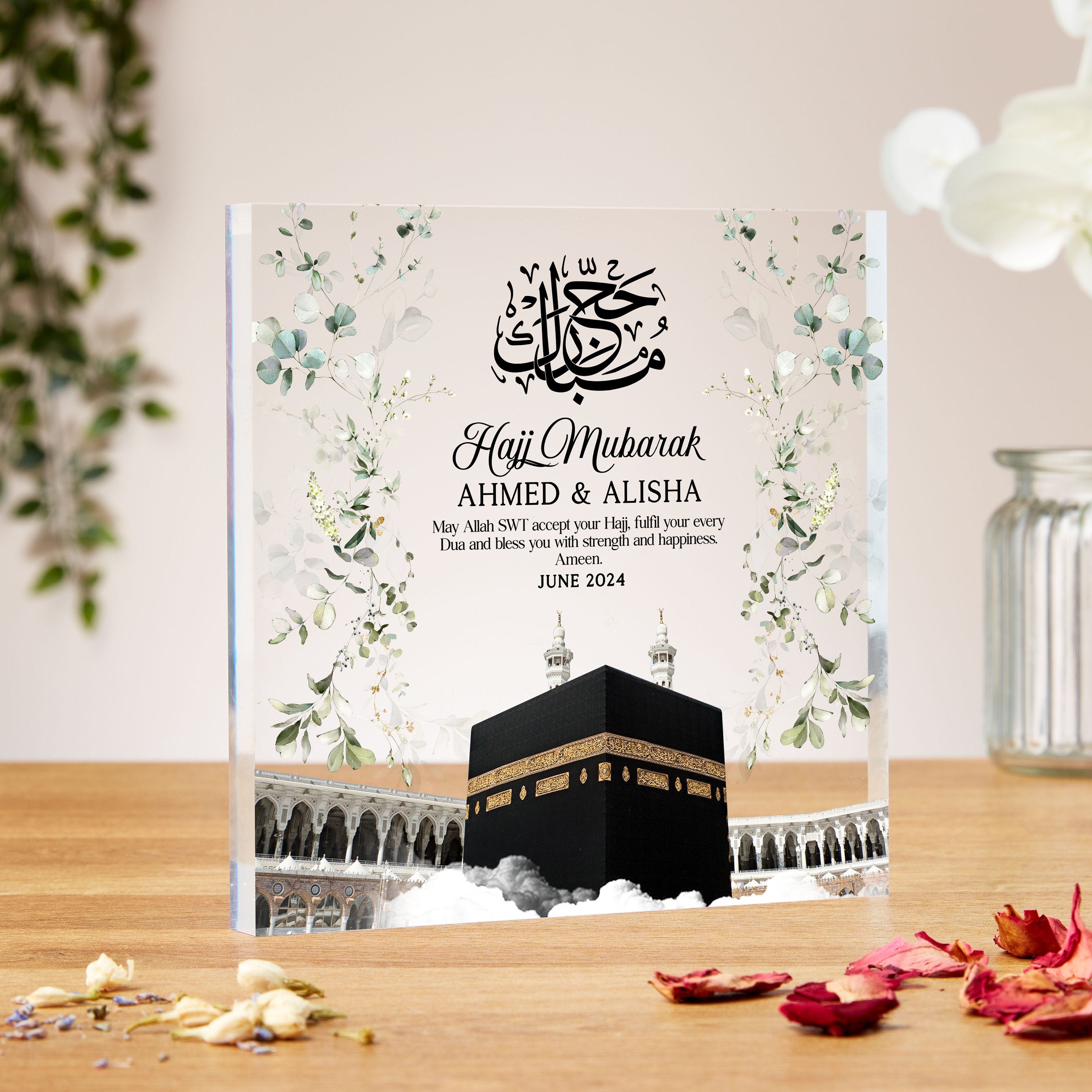 A crystal-clear acrylic plaque with soft floral decorations on the sides, featuring a detailed illustration of the Kaaba at the center with surrounding Masjid al-Haram architecture and clouds at the base. Above the Kaaba is Arabic calligraphy and a message that reads: “Hajj Mubarak – AHMED & ALISHA. May Allah SWT accept your Hajj, fulfil your every Dua and bless you with strength and happiness. Ameen.” The date “JUNE 2024” appears beneath the message. The design combines elegance and spirituality with a ser
