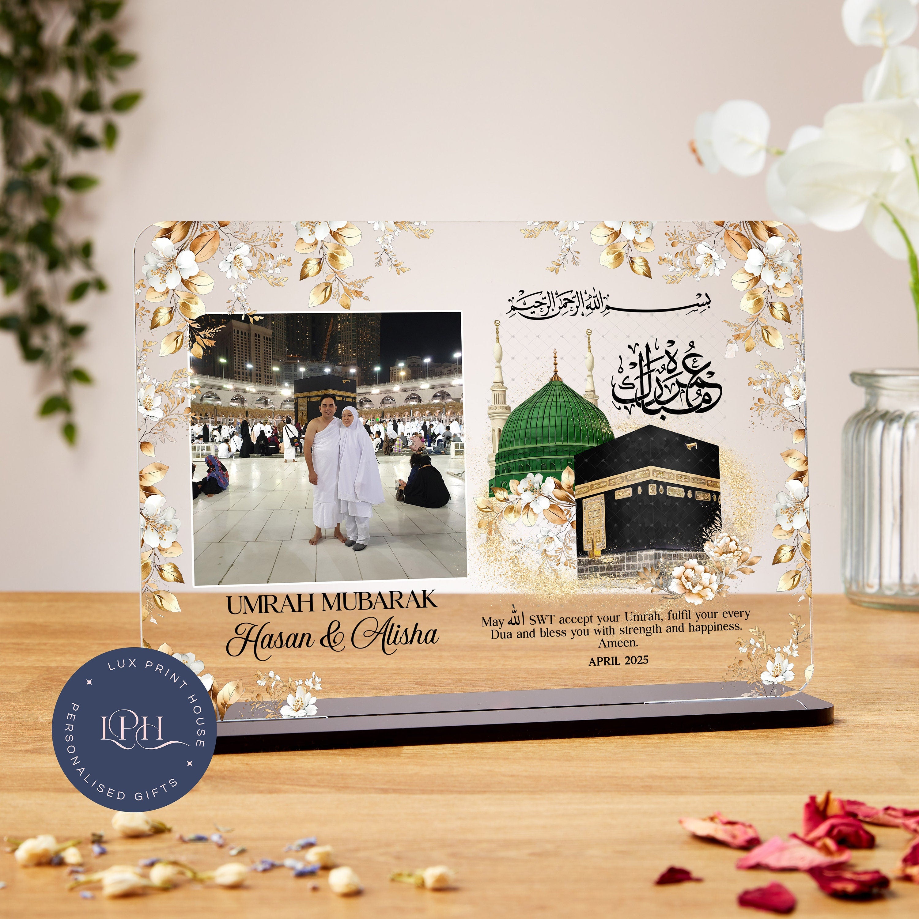 A personalized Umrah plaque decorated with golden floral designs and featuring a real photograph of a couple, Hasan and Alisha, standing together in Ihram attire at the Masjid al-Haram with the Kaaba in the background. To the right of the photo is an illustration of the Green Dome of the Prophet's Mosque and the Kaaba, along with Arabic calligraphy. The message reads: “UMRAH MUBARAK – Hasan & Alisha. May Allah SWT accept your Umrah, fulfil your every Dua and bless you with strength and happiness. Ameen.” Th