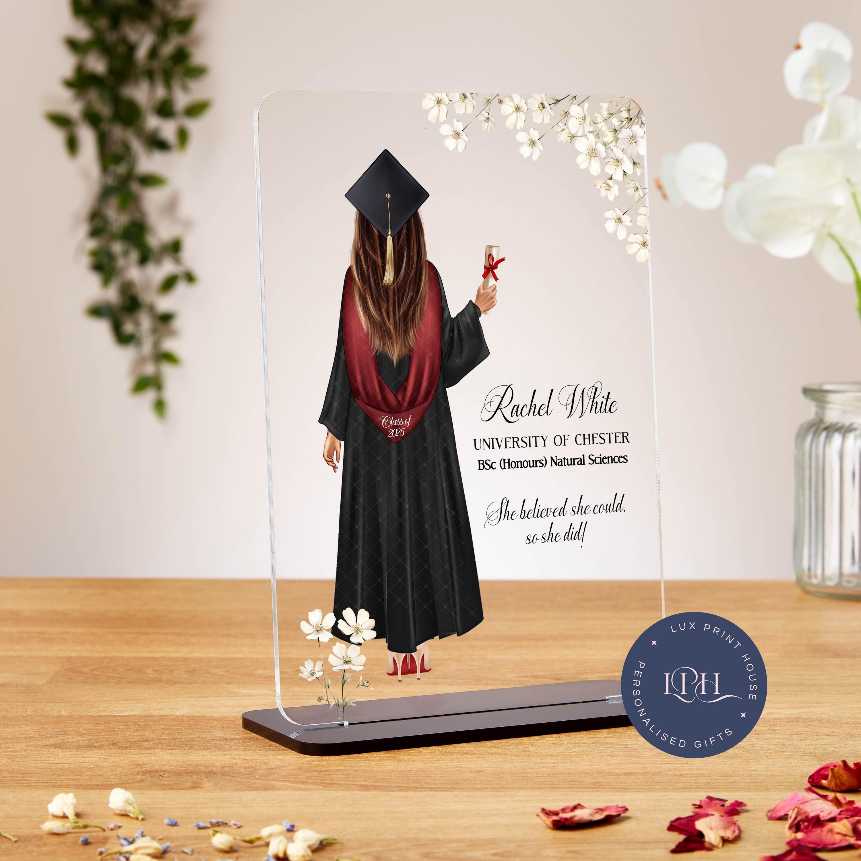A custom graduation plaque featuring an illustrated female graduate with long brown hair, wearing a black gown, maroon stole labeled “Class of 2025,” and a graduation cap with a gold tassel. She holds a diploma tied with a red ribbon. White floral accents decorate the corners. Text reads: “Rachel White, University of Chester, BSc (Honours) Natural Sciences. She believed she could, so she did!” A circular Lux Print House logo appears at the bottom.