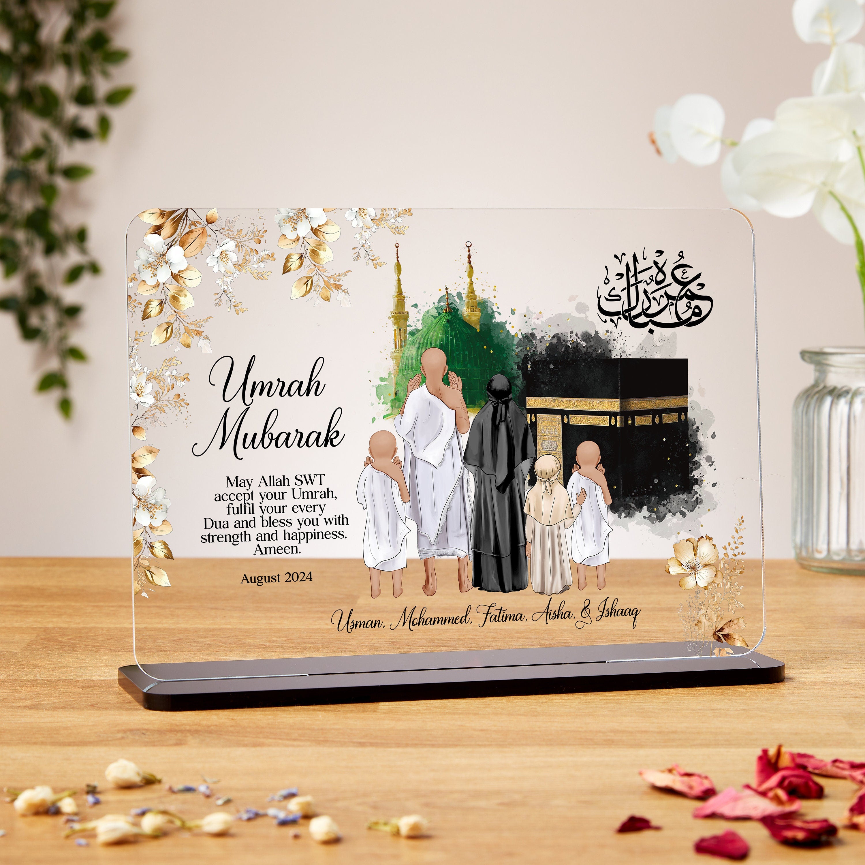 A decorative plaque with floral borders featuring an illustration of a family of five dressed in traditional Umrah attire, standing in front of the Kaaba and the Green Dome of the Prophet's Mosque. The text reads: “Umrah Mubarak. May Allah SWT accept your Umrah, fulfil your every Dua and bless you with strength and happiness. Ameen.” Below the message, it says "August 2024" and includes names: Usman, Mohammed, Fatima, Aisha, & Ishaaq. Arabic calligraphy is displayed in the top right corner.