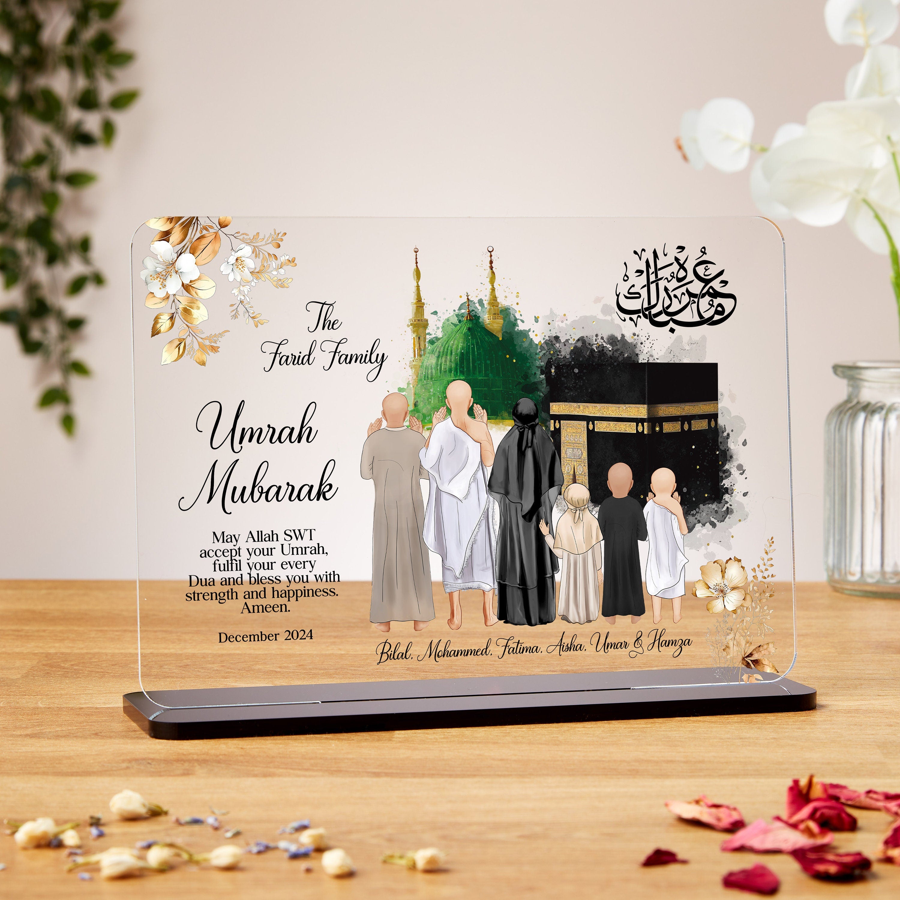 Alt text: A decorative glass plaque with floral designs in the corners, showing an illustrated family of seven in Umrah attire standing in front of the Kaaba and the Green Dome of the Prophet's Mosque. The text reads: “The Farid Family – Umrah Mubarak. May Allah SWT accept your Umrah, fulfil your every Dua and bless you with strength and happiness. Ameen.” The date “December 2024” appears below the prayer, followed by the names: Bilal, Mohammed, Fatima, Aisha, Umar & Hamza. Arabic calligraphy is displayed i