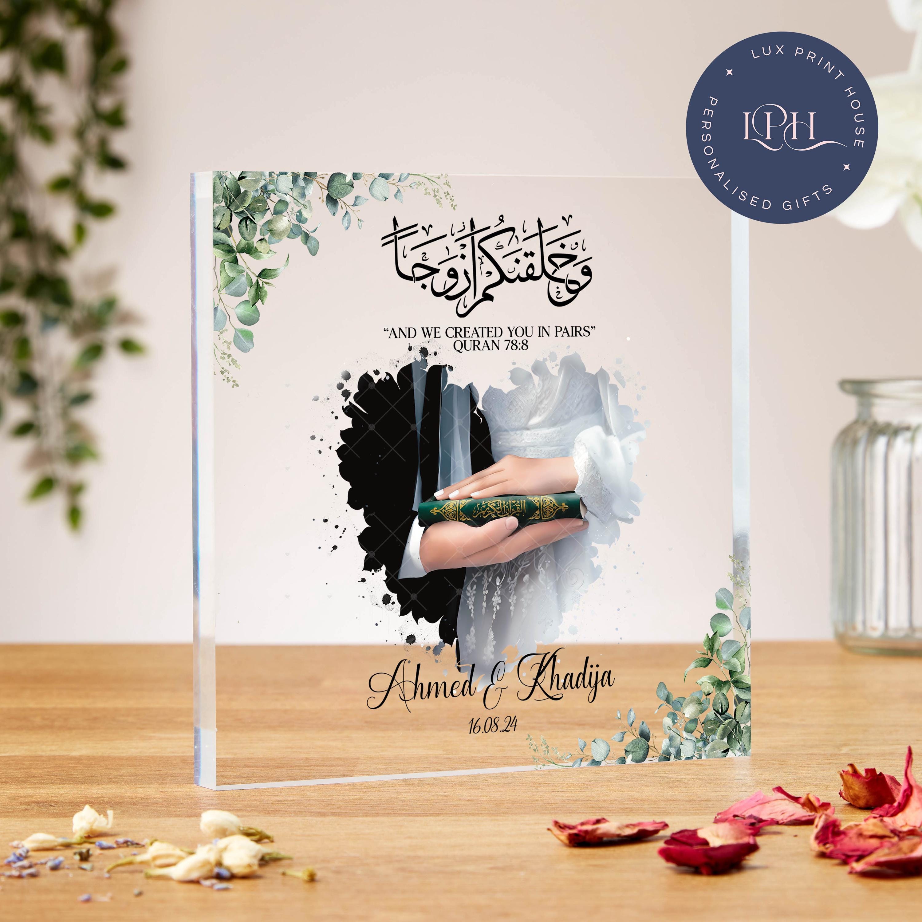 A romantic wedding-themed acrylic plaque with floral greenery in the corners. At the center is an illustration of a bride and groom’s hands holding a Quran together, with the bride in a white gown and the groom in black. A heart-shaped background blends into the design. Arabic calligraphy at the top is paired with the Quran verse: “And We created you in pairs” (Quran 78:8). Below, the names “Ahmed & Khadija” and the date “16.08.24” are elegantly written.